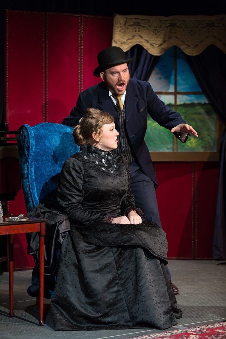 Tom Sitzler, dressed in a bowler hat and period waistcoat, conversing with a woman in a black floor-length gown in "The Boor."