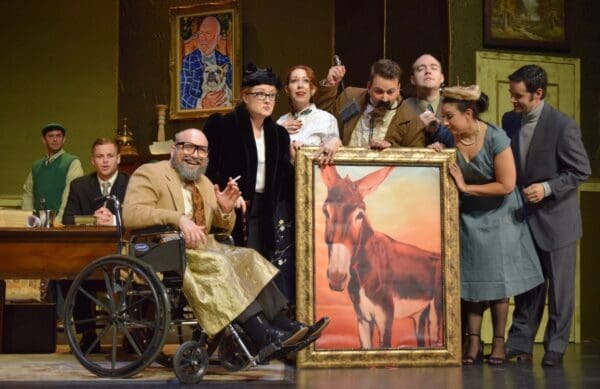 Tom Sitzler as Gianni Schicchi, sitting in a wheelchair surrounded by the rest of the cast as they hold up a large painting of a donkey.